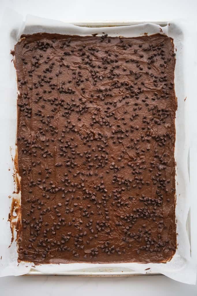 Chocolate mixture spread out thinly on parchment paper that is sitting on top of a baking pan, topped with chocolate chips