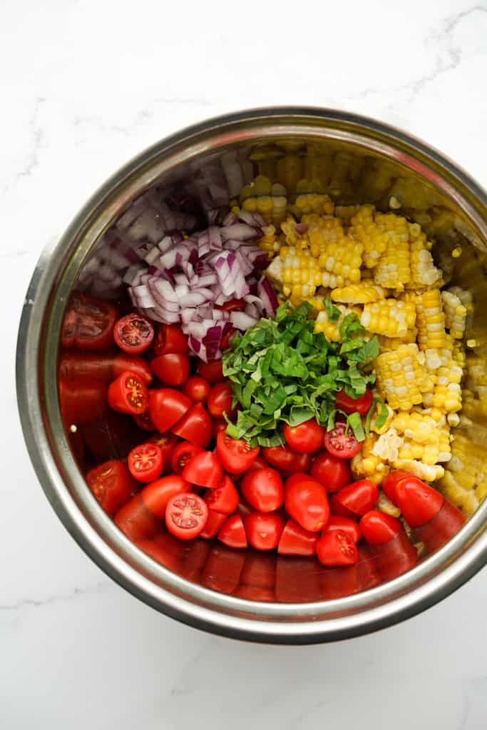 Top down view of a mixing bowl with corn kernels, halved cherry tomatoes, diced red onions and fresh basil.