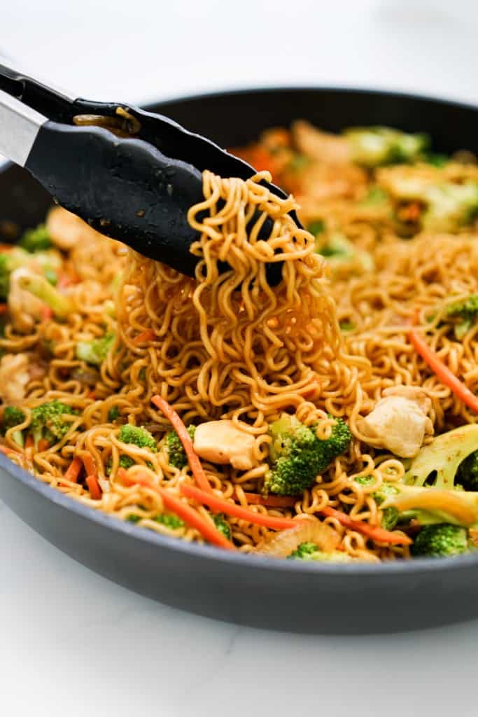 Using a pair of tongs to lift up ramen noodles from a skillet