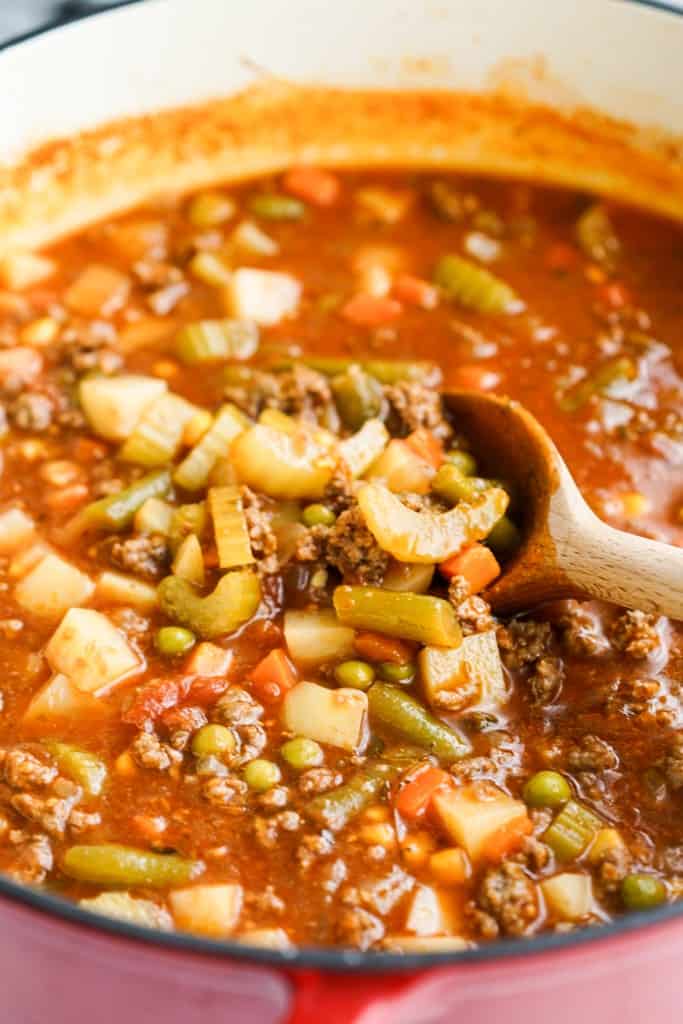 Wooden spoon scooping up a large pot of soup loaded with ground beef and veggies