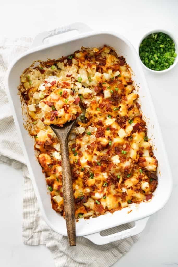 A brown spatula digging to a casserole dish loaded with creamy potato, bacon, cheese and green onions