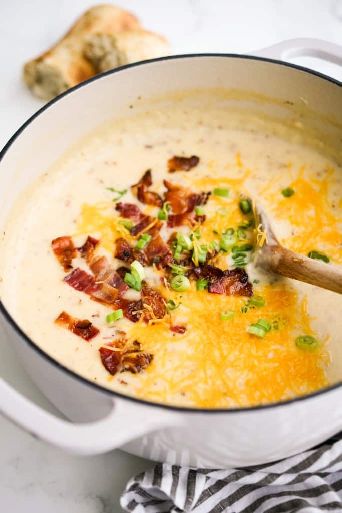 Wooden spoon stirring a pot of mashed potato soup topped with bacon, cheese and chopped green onions