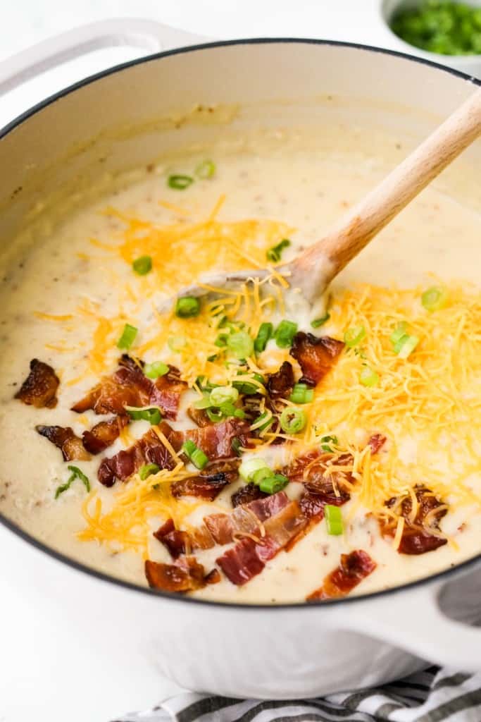 A pot of mashed potato soup topped with bacon pieces, shredded cheddar cheese and green onions, and a wooden spoon in the soup