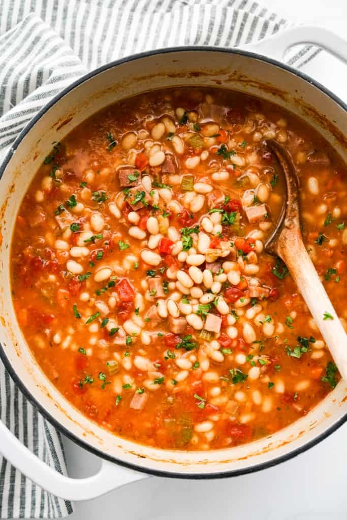 A top down view of a large pot of navy bean soup with ham, veggies and herbs. 