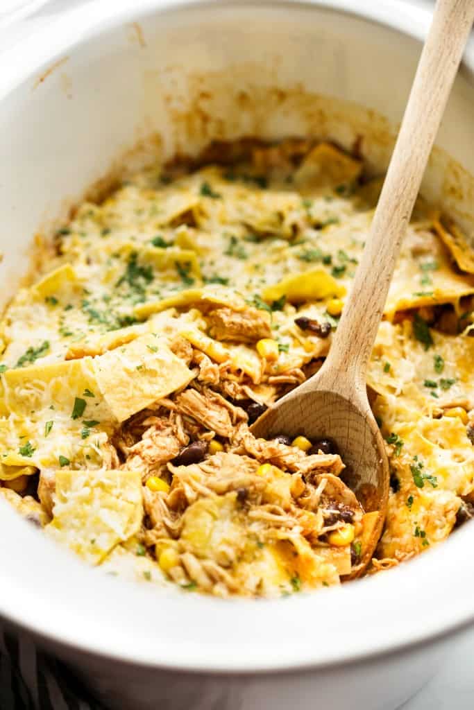 A wooden spoon digging into a crockpot with a mixture of shredded chicken, tortillas and cheese