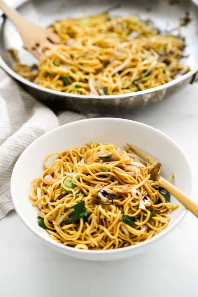 A bowl of vegetable chow mein with a large skillet of it in the background