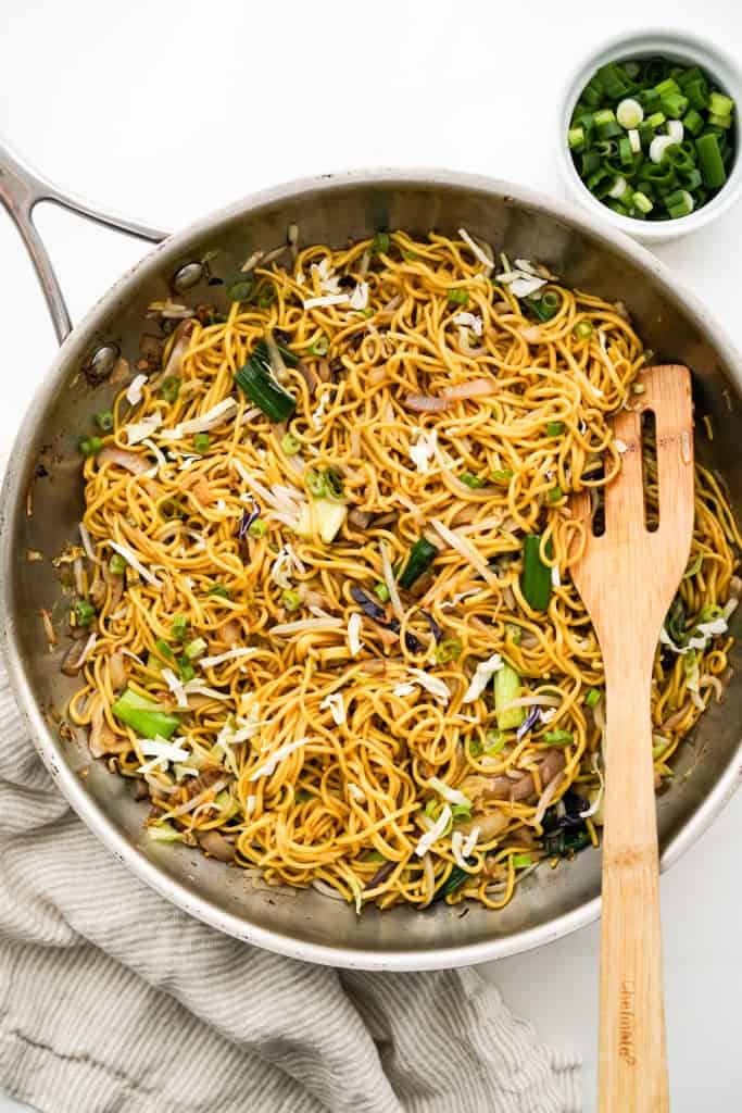 A skillet of vegetable chow mein with a wooden spatula in it.