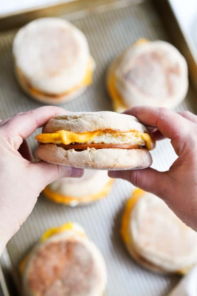 Hands holding a english muffin breakfast sandwich, with more in the background