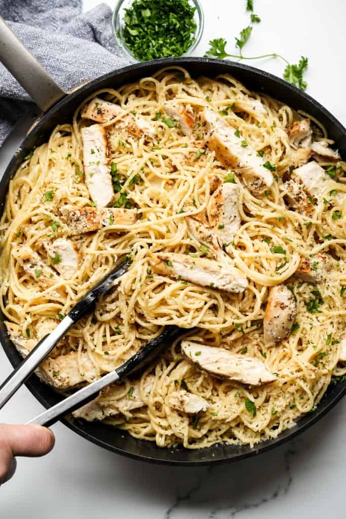 a pair of tongs gripping onto garlic parmesan spaghetti noodles in a skillet topped with chicken