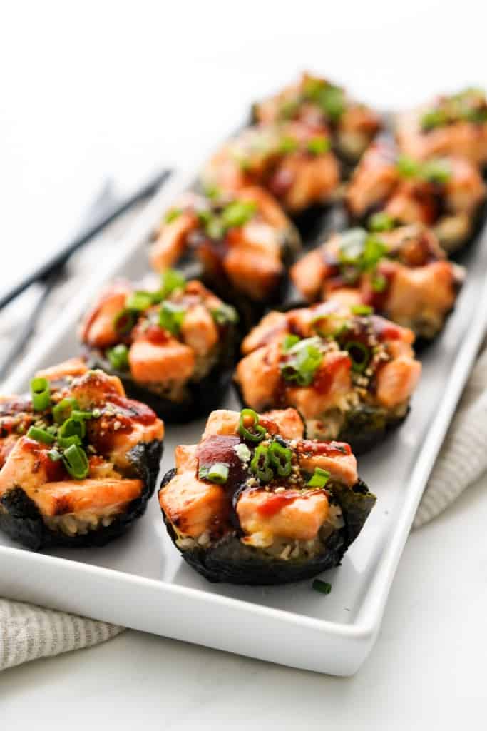 A rectangular plate of salmon sushi cups topped with chopped green onions, sesame seeds and drizzled with sauces.
