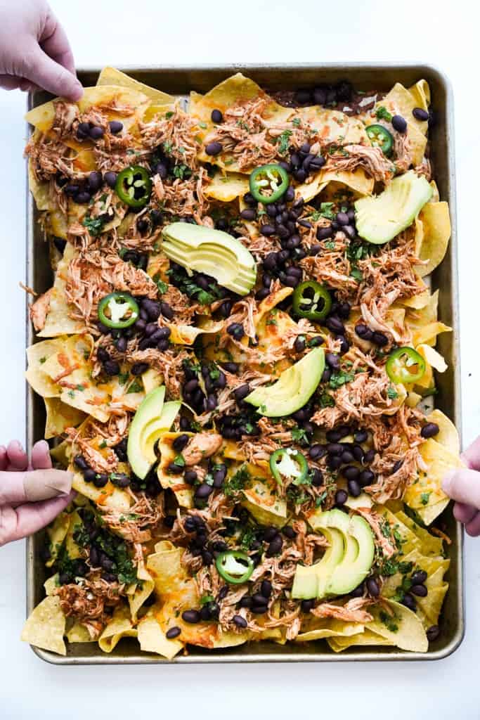 a few hands picking up tortilla chips from shredded chicken nachos on a sheet pan