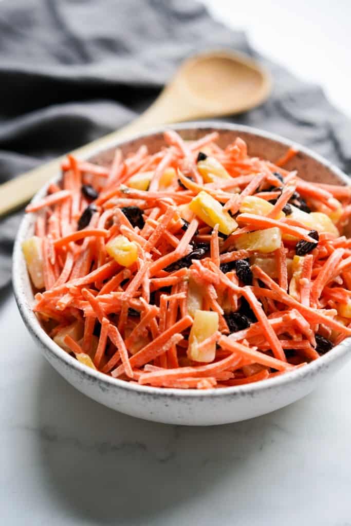 A bowl of carrot raisin pineapple salad, with a wooden spoon in the background