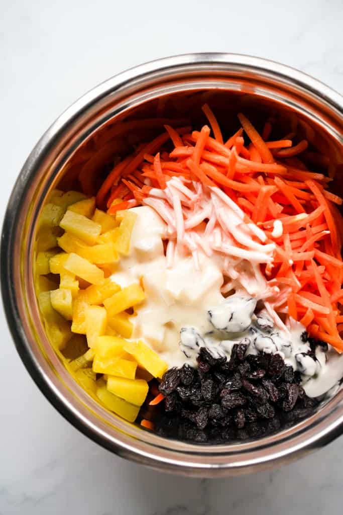 Mayo dressing added to a bowl of shredded carrots, pineapple and raisins