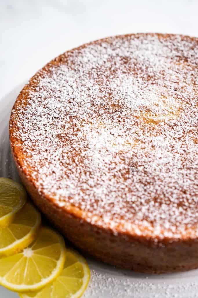 Whole lemon olive oil round cake on a cake stand topped with powdered sugar, with lemon slices on the side.