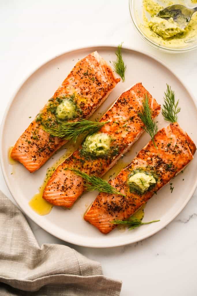 Top down view of a round plate of three salmon fillets topped with dill garlic butter compound