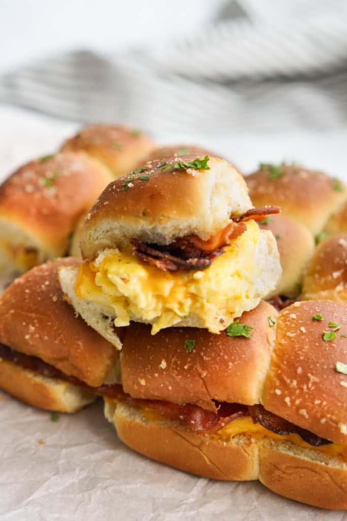 A Hawaiian roll breakfast slider sitting on top of a few rows of sliders, loaded with scrambled eggs and bacon