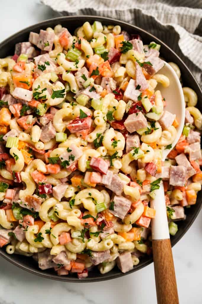 Digging into a bowl of macaroni ham pasta salad with a white spoon