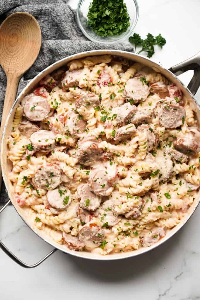 A large pot of rotini pasta and smoked sausage tossed in creamy white sauce. A wooden ladle on the side
