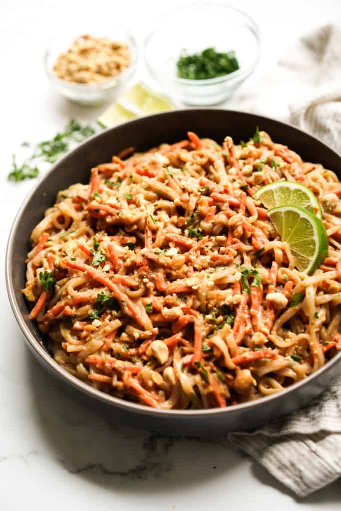 A bowl of rice noodles tossed in peanut sauce, topped with shredded carrots, peanuts and lime wedges