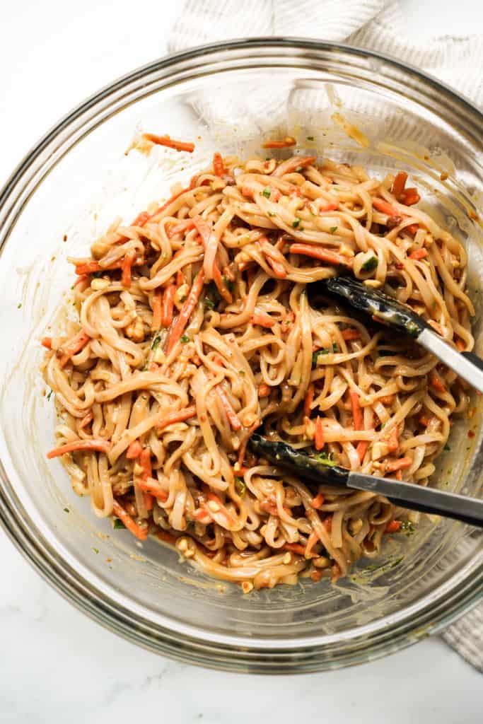 rice noodles and vegetables tossed in thick peanut sauce with a pair of tongs