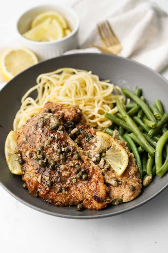 A bowl with two pieces of lemon chicken piccata served with pasta and green beans