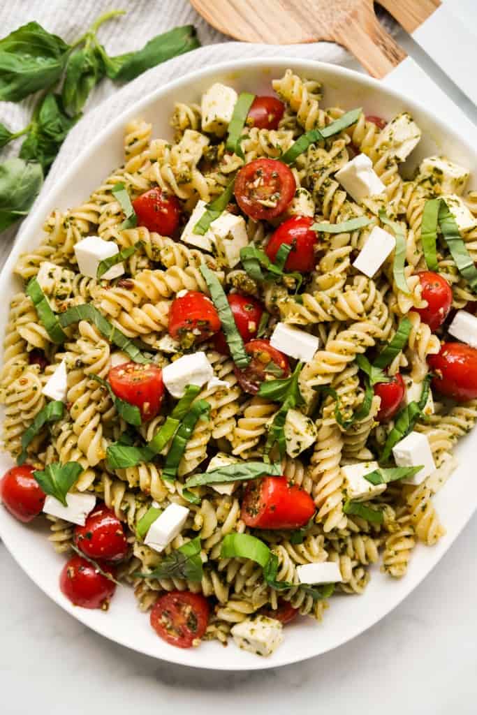 A large bowl of pesto pasta salad tossed with cubed feta, cherry tomatoes and fresh basil