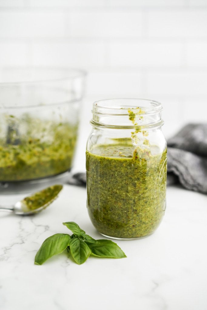 A jar of pistachio pesto with basil leaves on the side.