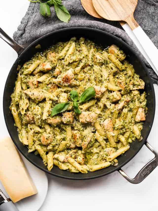 A large skillet loaded with creamy pesto pasta and chicken pieces, with fresh basil leaves and a block of parmesan cheese in the background
