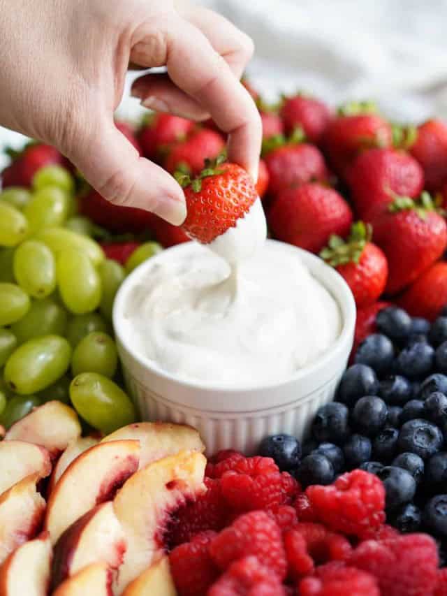 Dipping a strawberries into a bowl of fruit dip, with fruits surrounding it.