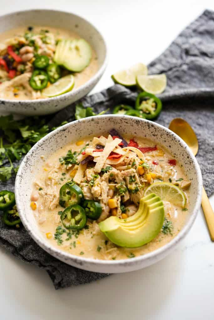 Two bowls of white chicken chili garnished with sliced avocado, cilantro and tortilla chips