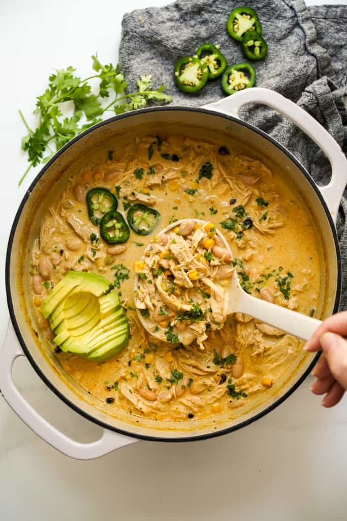 A large pot of creamy white chicken chili topped with jalapeño, sliced avocados and cilantro.