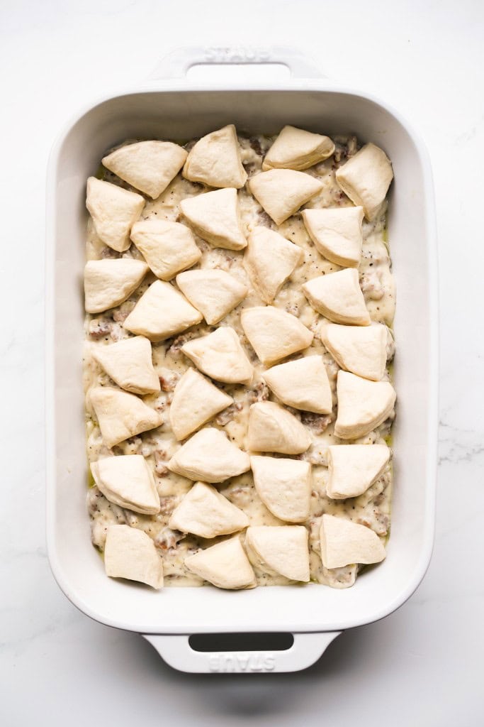 rectangular casserole is filled with thick sausage gravy topped with small bite size pieces of raw biscuit dough