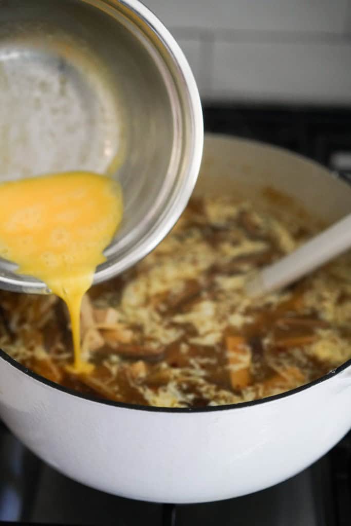 Pouring whisked eggs into a large pot of soup on the stove top
