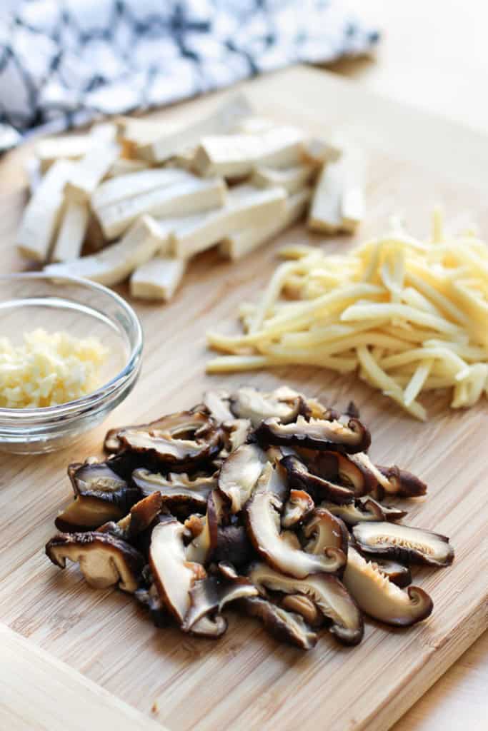 Minced garlic, tofu slices, bamboo shoot strips and sliced shiitake mushrooms on a cutting board