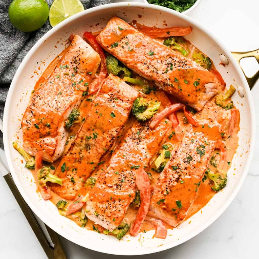 A large skillet with salmon fillets coated in red curry sauce, with broccoli and red bell peppers in the sauce.