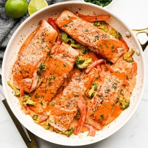 A large skillet with salmon fillets coated in red curry sauce, with broccoli and red bell peppers in the sauce.