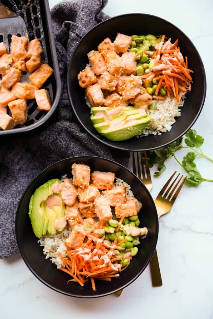 Two bowls of salmon bowl with rice, and veggies, with an air fryer filled with salmon bites on the side.