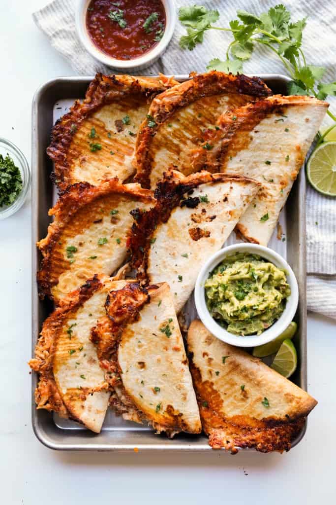 A large baking sheet filled with baked chicken tacos, with salsa and guacamole on the side