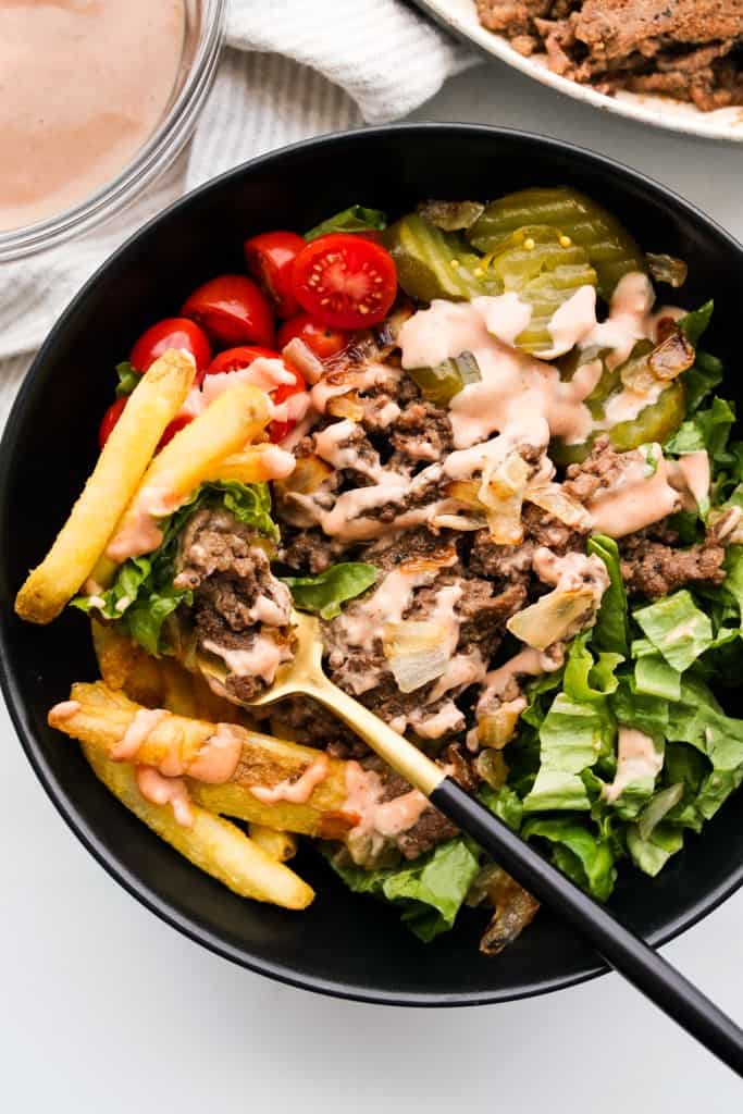 Digging into a burger bowl that is filled with lettuce, tomatoes, pickles and fries with a fork