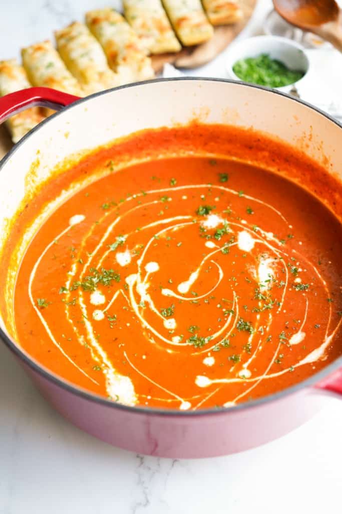 A pot of tomato bell pepper soup with cream drizzled on top, with garlic bread in the background