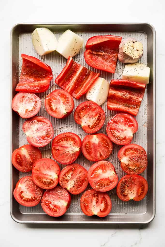 A large sheet pan with halved tomatoes, red bell peppers, onions and a bulb of garlic tossed with salt and black pepper
