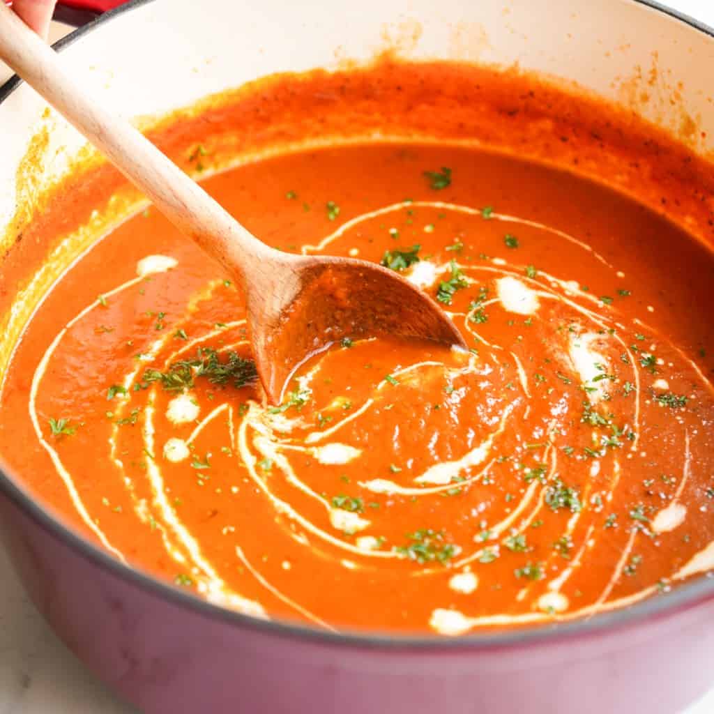 A Dutch oven filled with roasted red pepper and tomato soup, drizzled with heavy cream