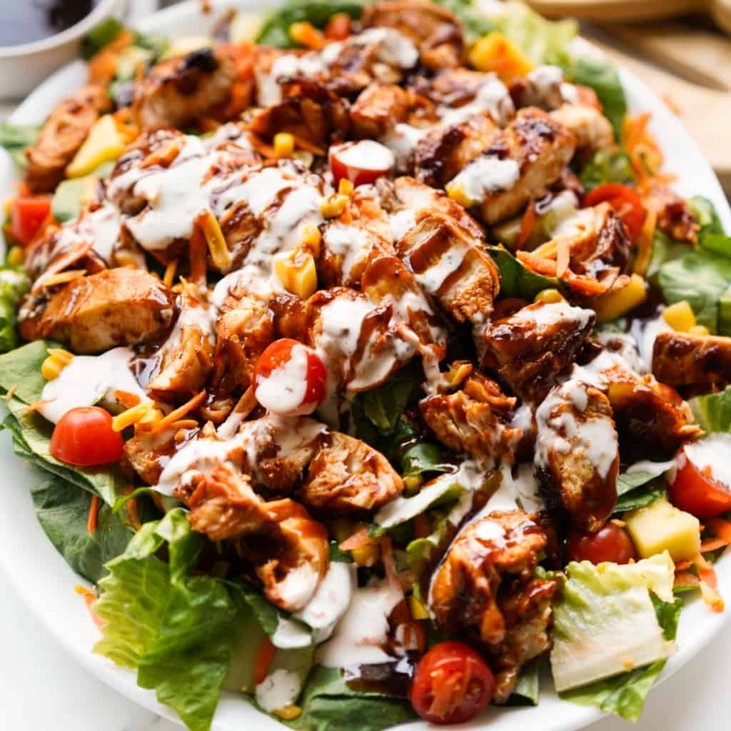 A platter piled with green lettuce, tomatoes, cheese and chunks of bbq chicken, drizzled with ranch and bbq sauce