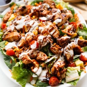 A platter piled with green lettuce, tomatoes, cheese and chunks of bbq chicken, drizzled with ranch and bbq sauce