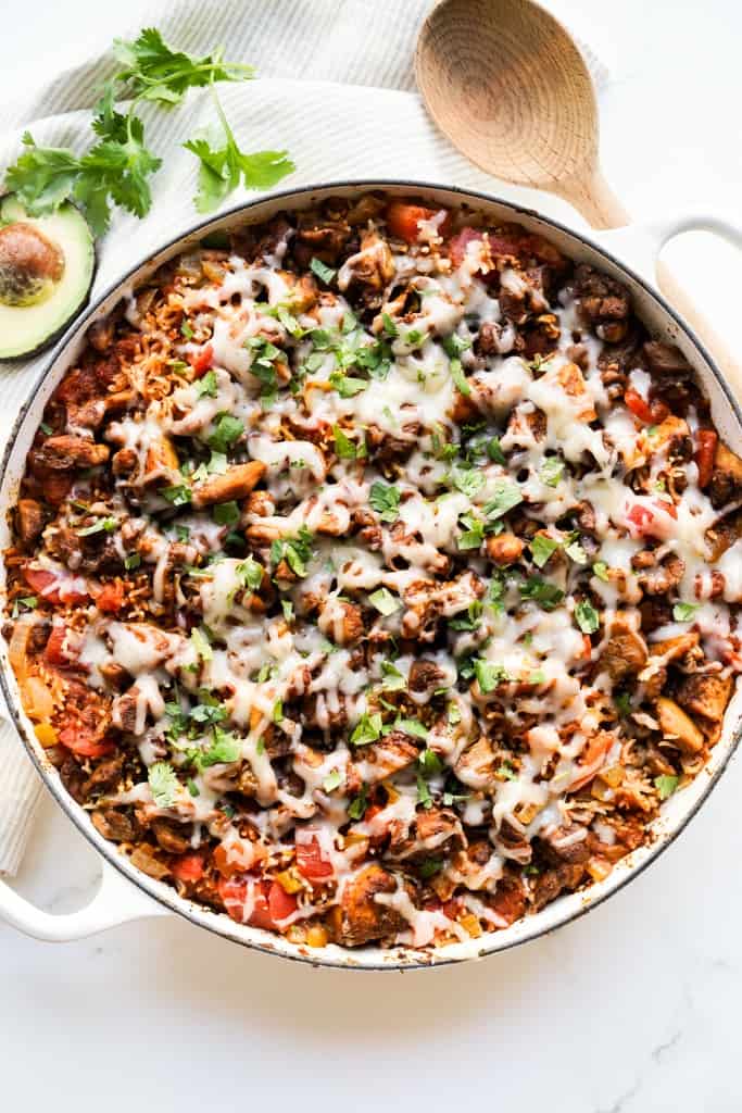 Top down view of a one pot Mexican chicken and rice dish in a braiser, with a wooden spoon, avocado and cilantro in the background
