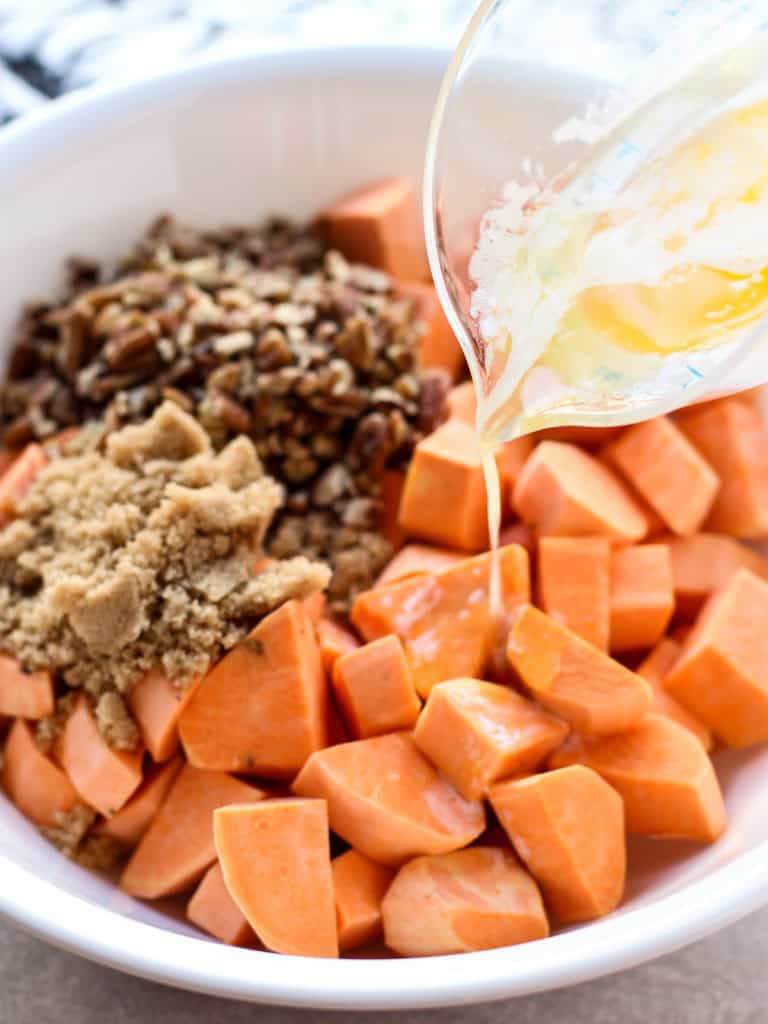 Pouring melted butter into a bowl loaded with cubed sweet potatoes, chopped pecans and brown sugar