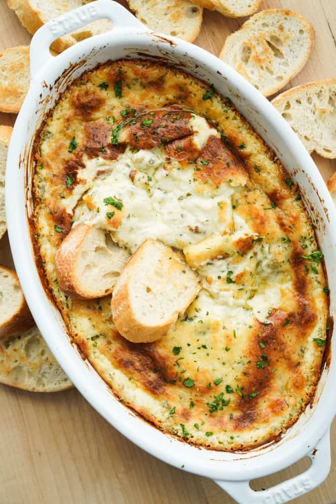 A oval dish of warm roasted garlic dip with baguette dipped in it and more baguettes around it
