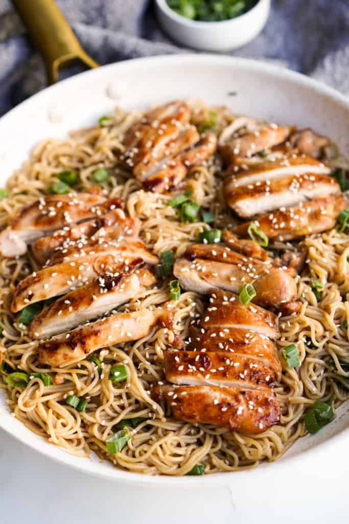 Sliced teriyaki chicken placed on top of ramen noodles in a skillet, garnished with green onions and sesame seeds