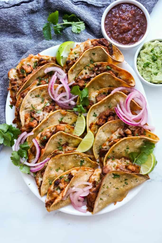 A platter loaded with crispy baked chipotle chicken tacos stuffed with cheese, with salsa and guacamole on the side