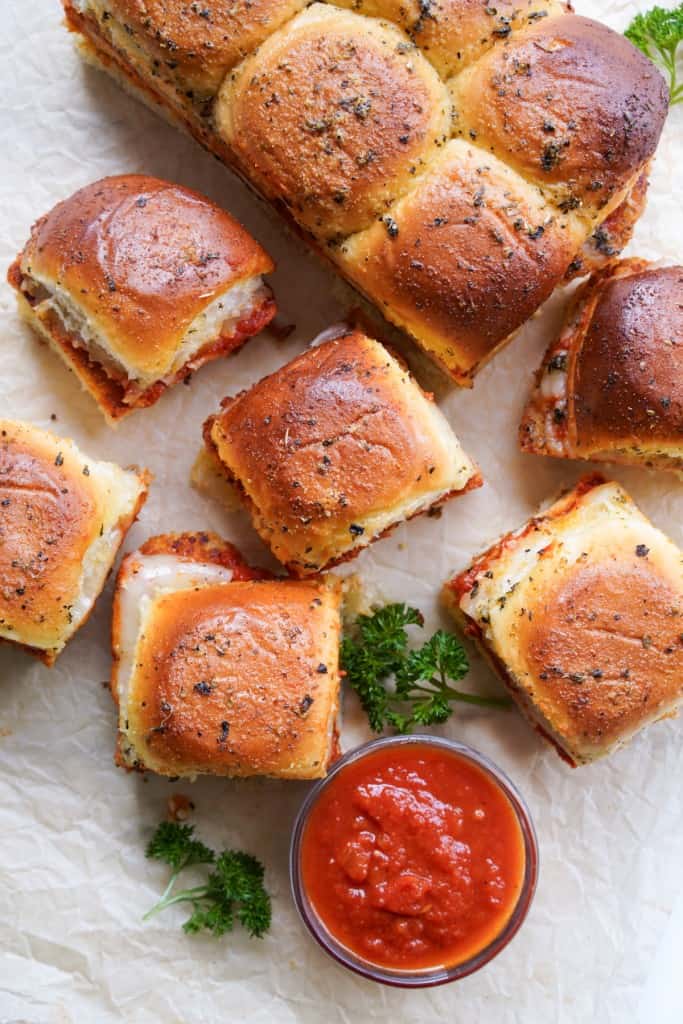 A top down picture showing the lovely browned tops of several Italian chicken sliders accented with fresh parsley and a small bowl of marinara sauce.